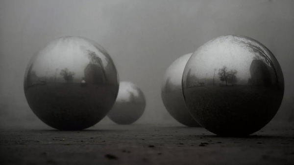 Reflective metallic spheres resting on the ground enveloped in heavy fog, symbolizing the coexistence of contemporary art and heavy industry.