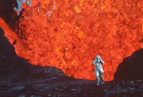 A volcanologist wearing a silver heat protective suit stands dwarfed in front of a colossal wall of erupting red lava.
