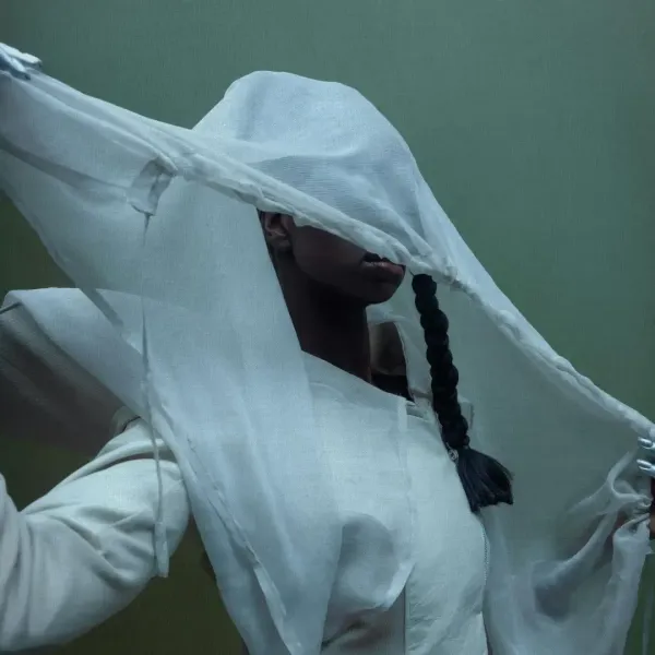 A model's face obscured by draped sheer white fabric against a muted green background, symbolizing the synthetic veil of modern textiles.