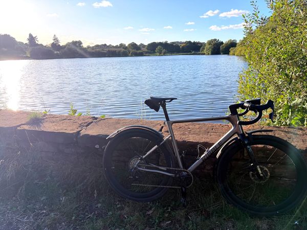 A bike leaning against a sandstone wall.  A lake in the background surrounded by trees.  It looks a beautiful day with only a few clouds int he sky and the sun reflecting on the water.