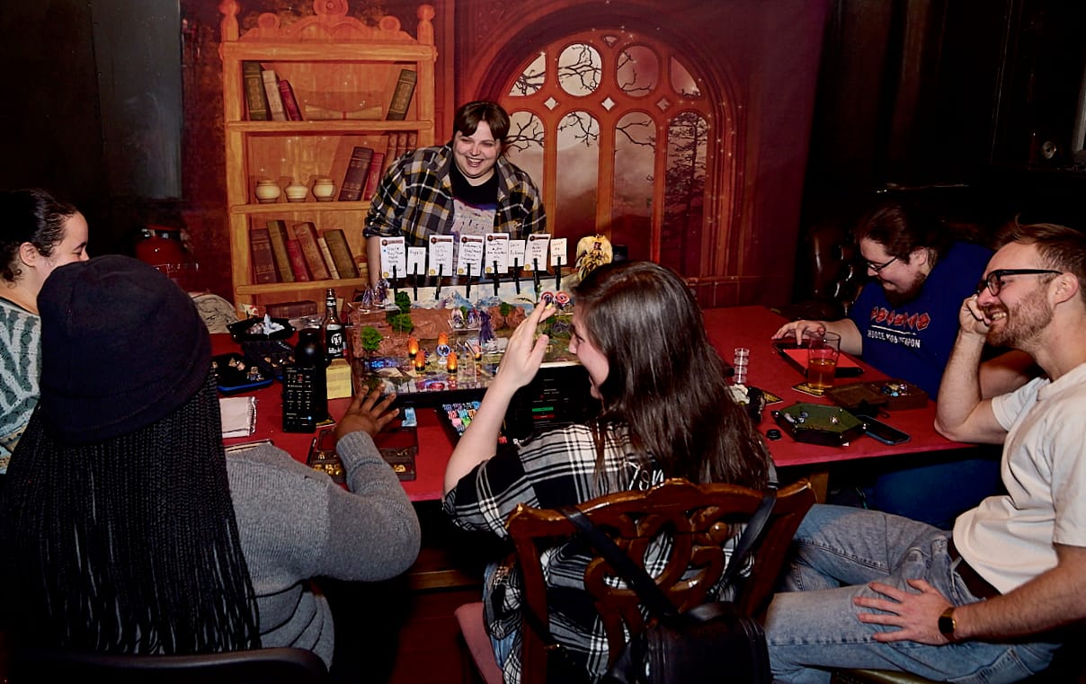 People gather around a table as a person runs a TTRPG before a painted backdrop.