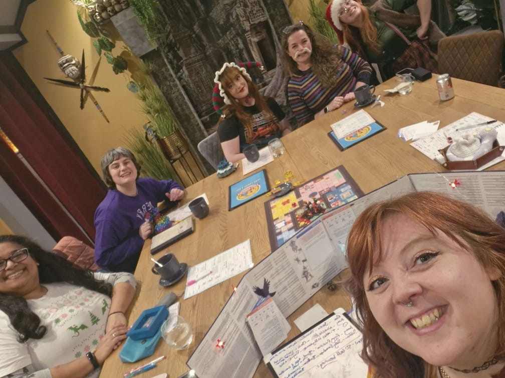 Several women smile and pose in a selfie taken around a table set up to play a TTRPG.