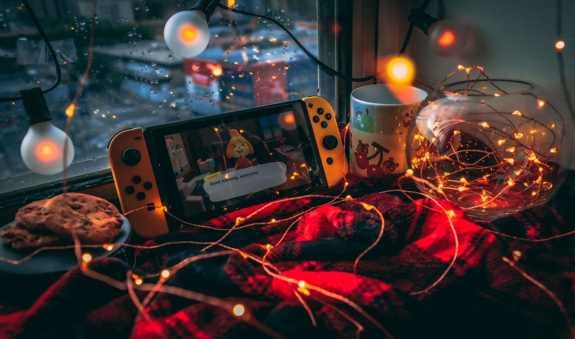 A Nintendo Switch open to Animal Crossing: New Horizons, sitting on a flannel blanket covered in fairy lights in front of a rainy windo