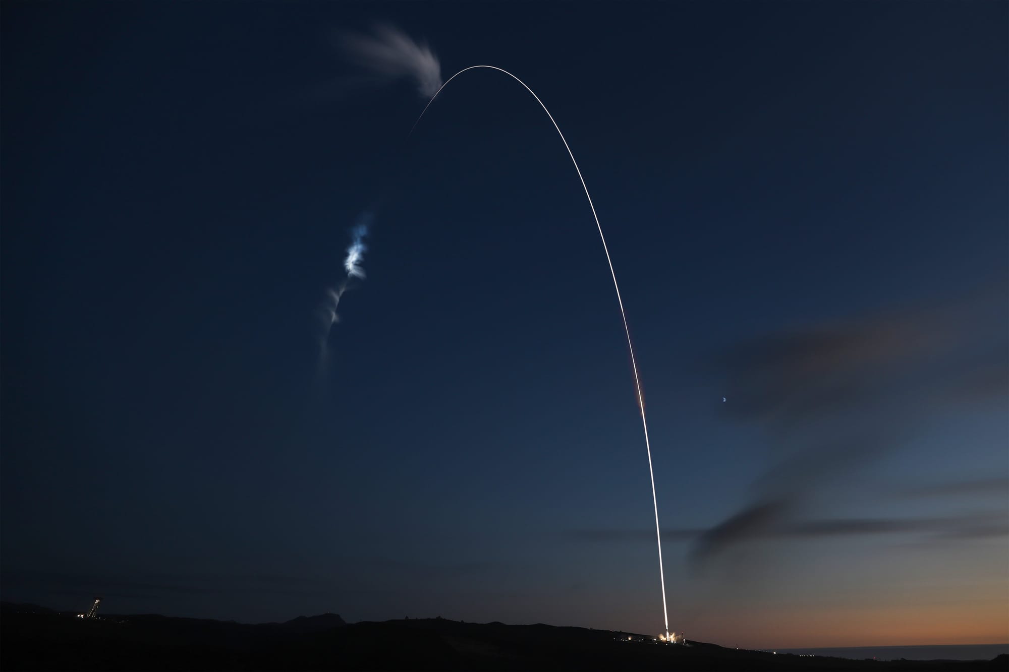 Long-exposure photo of a rocket launch at dusk, showing a bright arc of exhaust trail as the rocket ascends into space, with twilight skies and faint clouds illuminated above the launch site.