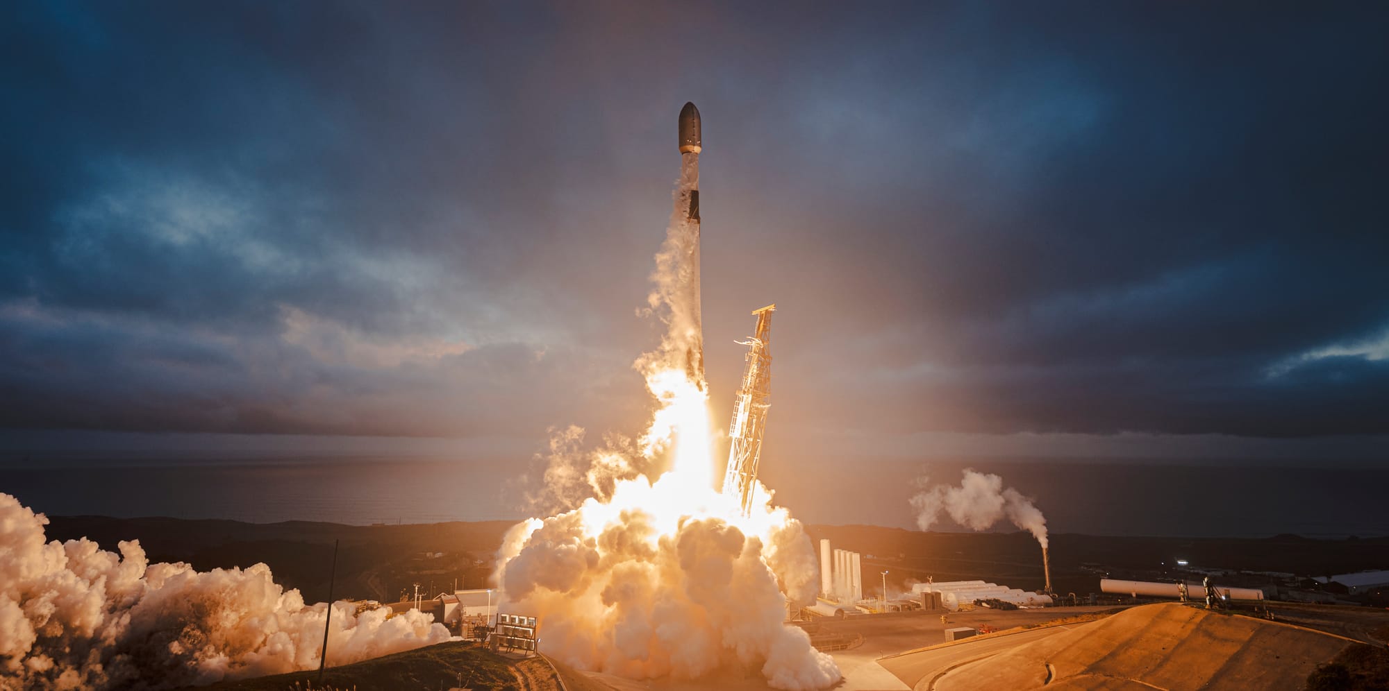 A Falcon 9 rocket launches at dawn from Vandenberg Space Force Base, engines blazing with fiery exhaust and billowing smoke as the vehicle ascends into the cloudy sky above.