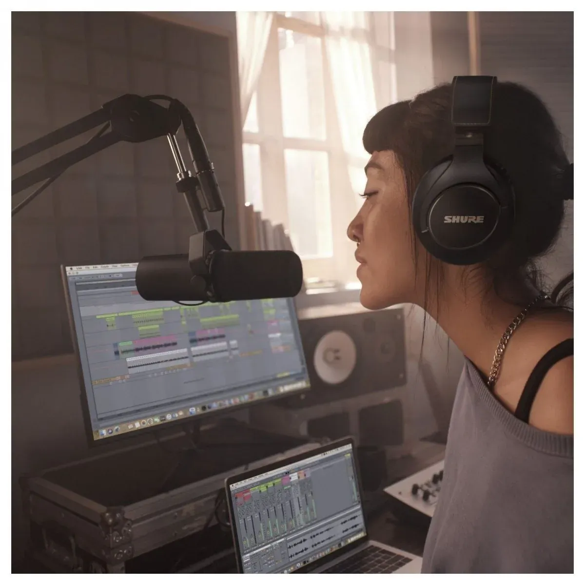 女生戴著監聽耳機,坐在窗邊的錄音桌前對著 Shure SM7B 錄音,旁邊的電腦開著錄音軟體