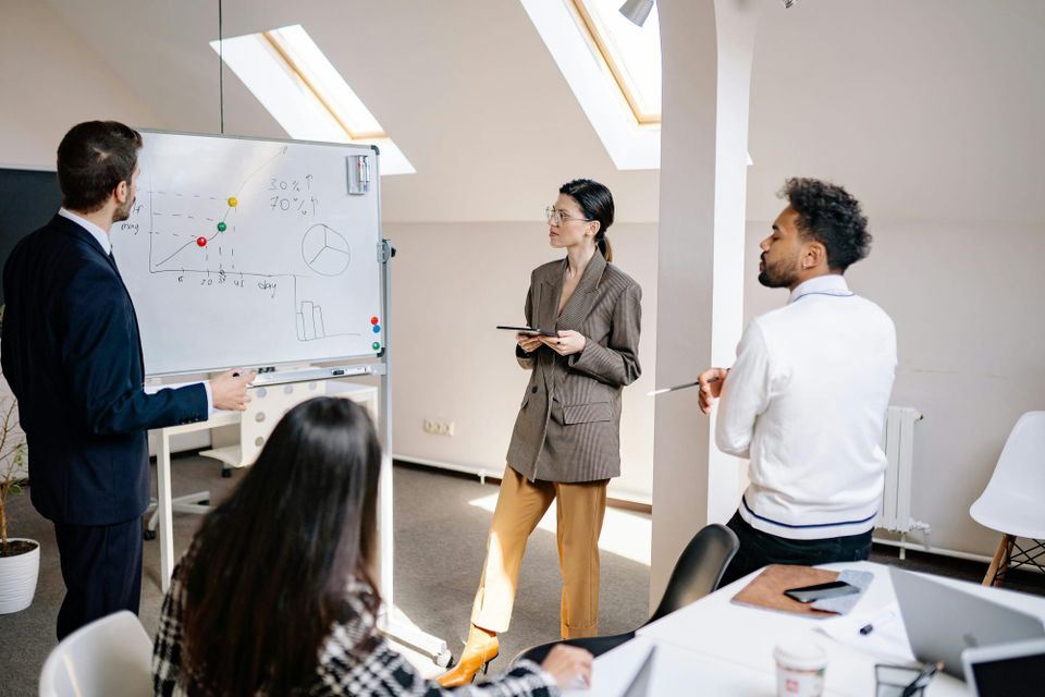 Image: Group of leaders look on at a whiteboard with a plotted line chart and pie graph drawn on it.