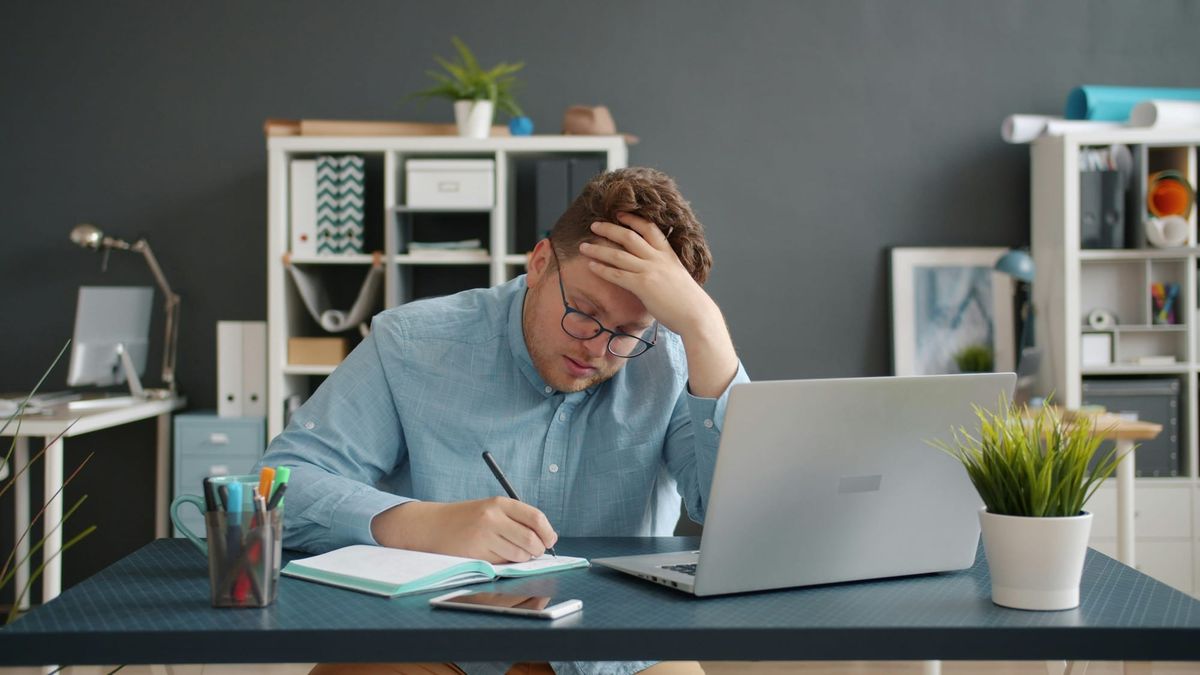A stressed man in glasses holds his head while writing at an office desk with a laptop nearby.
