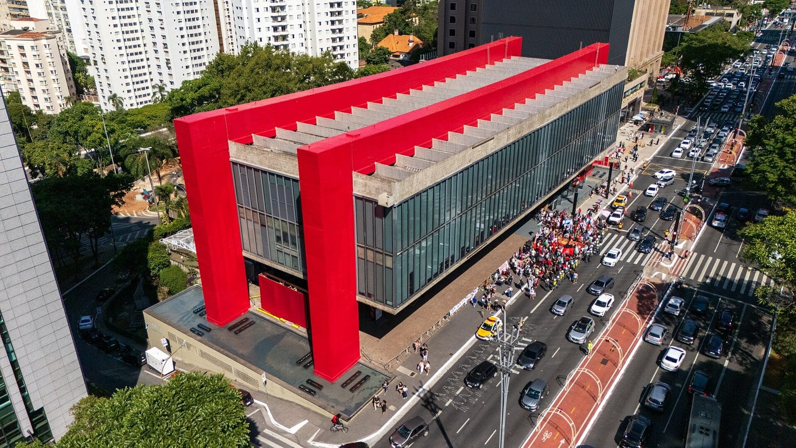Luftbild des MASP an der Avenida Paulista: ein Glasbau, der an zwei roten Betonportiken über einem offenen Platz hängt. Auf der Strasse davor eine Menschenmenge mit Fahnen.