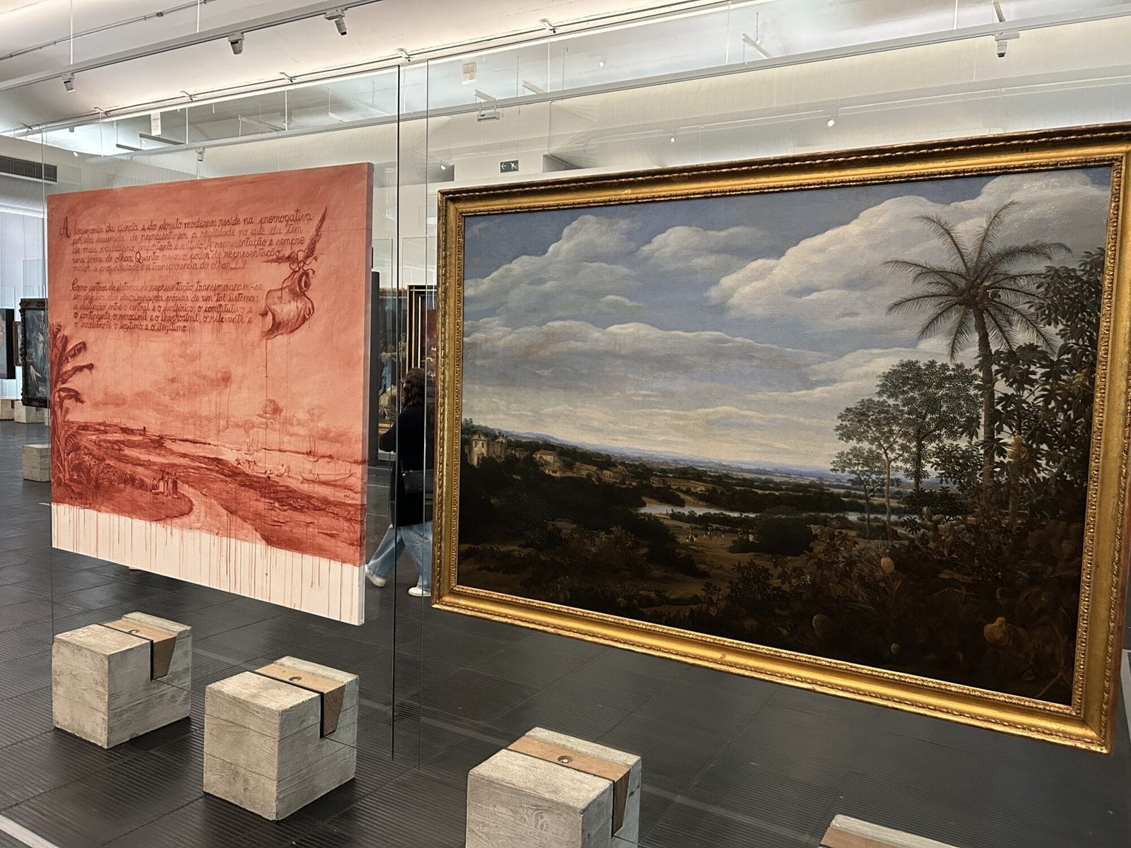 Links ein grossformatiges Bild in Rot mit handschriftlichem Text, rechts eine barocke brasilianische Landschaft mit Palmen in einem breiten Goldrahmen. Beide hängen frei im Raum.