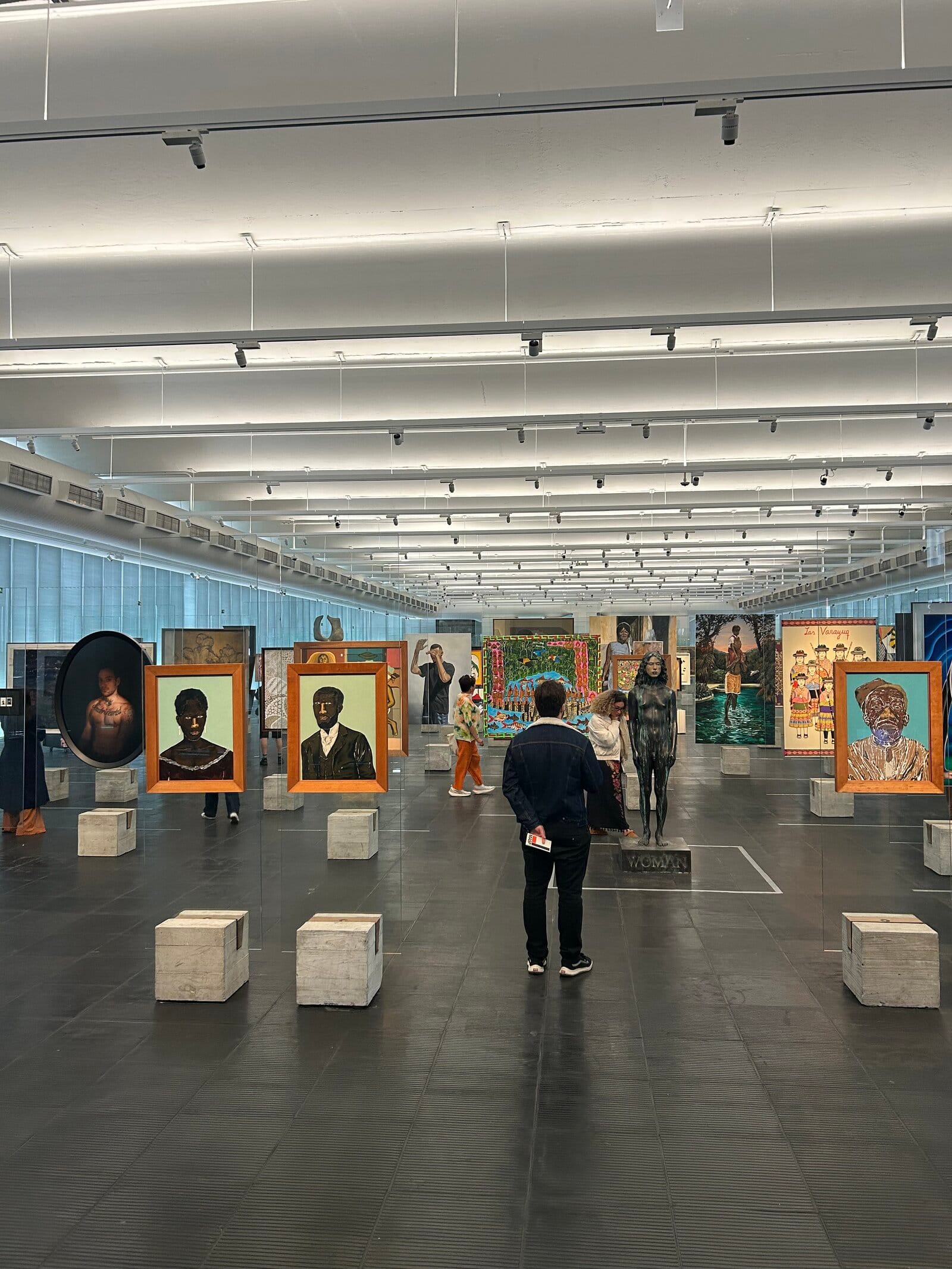 Blick in den Ausstellungssaal des MASP. Eine Person steht allein in der Mitte des leeren Bodens, im Hintergrund Gemälde auf Glasstelen und weitere Besucherinnen und Besucher.