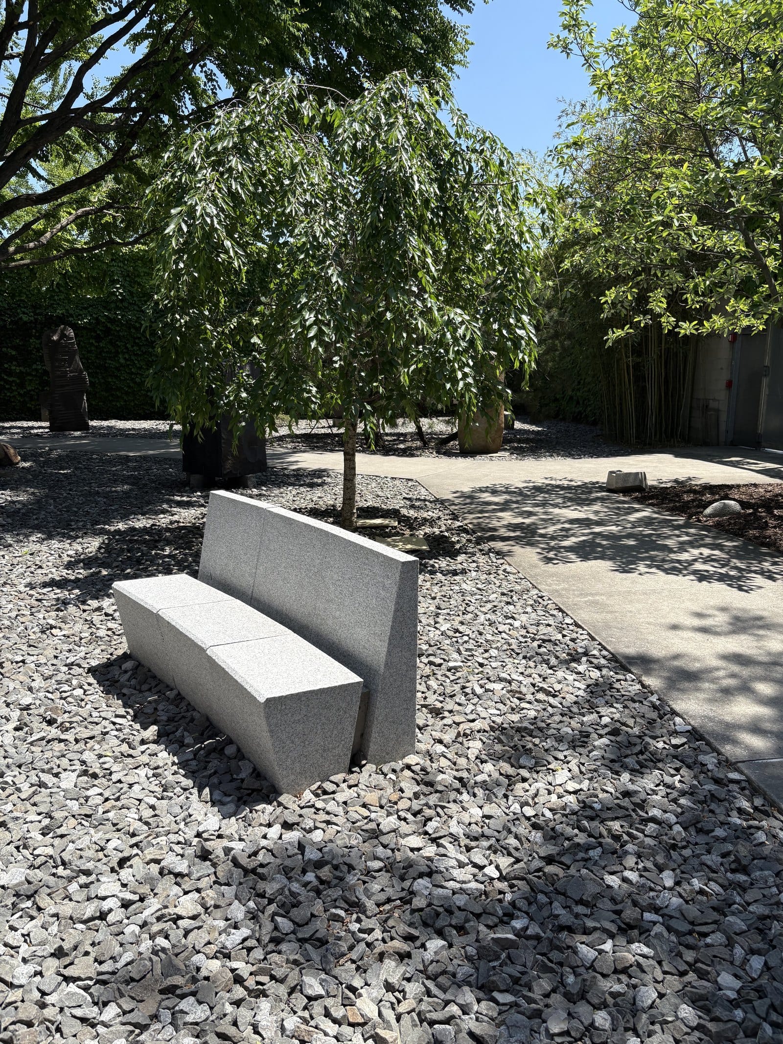 Eine helle Granitbank mit Rückenlehne steht im grauen Kies. Dahinter ein junger Baum, ein Betonweg und dunkle Skulpturen im Schatten.