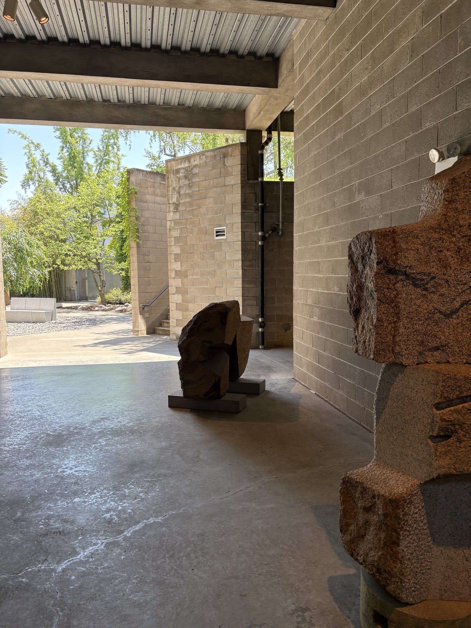 Blick aus einer überdachten Galerie mit Betonboden und Wänden aus Betonsteinen in den sonnigen Garten. Vorne rechts eine Skulptur aus rötlichem Granit, in der Mitte eine dunkle Steinskulptur auf einem Sockel, hinten links im Kies eine helle Steinbank.