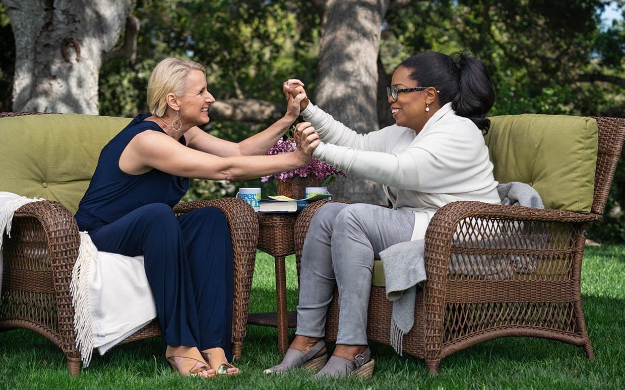 Elizabeth Gilbert and Oprah Winfrey on Super Soul Sunday. They're in lounge chairs on a grass lawn, clasping hands and smiling.