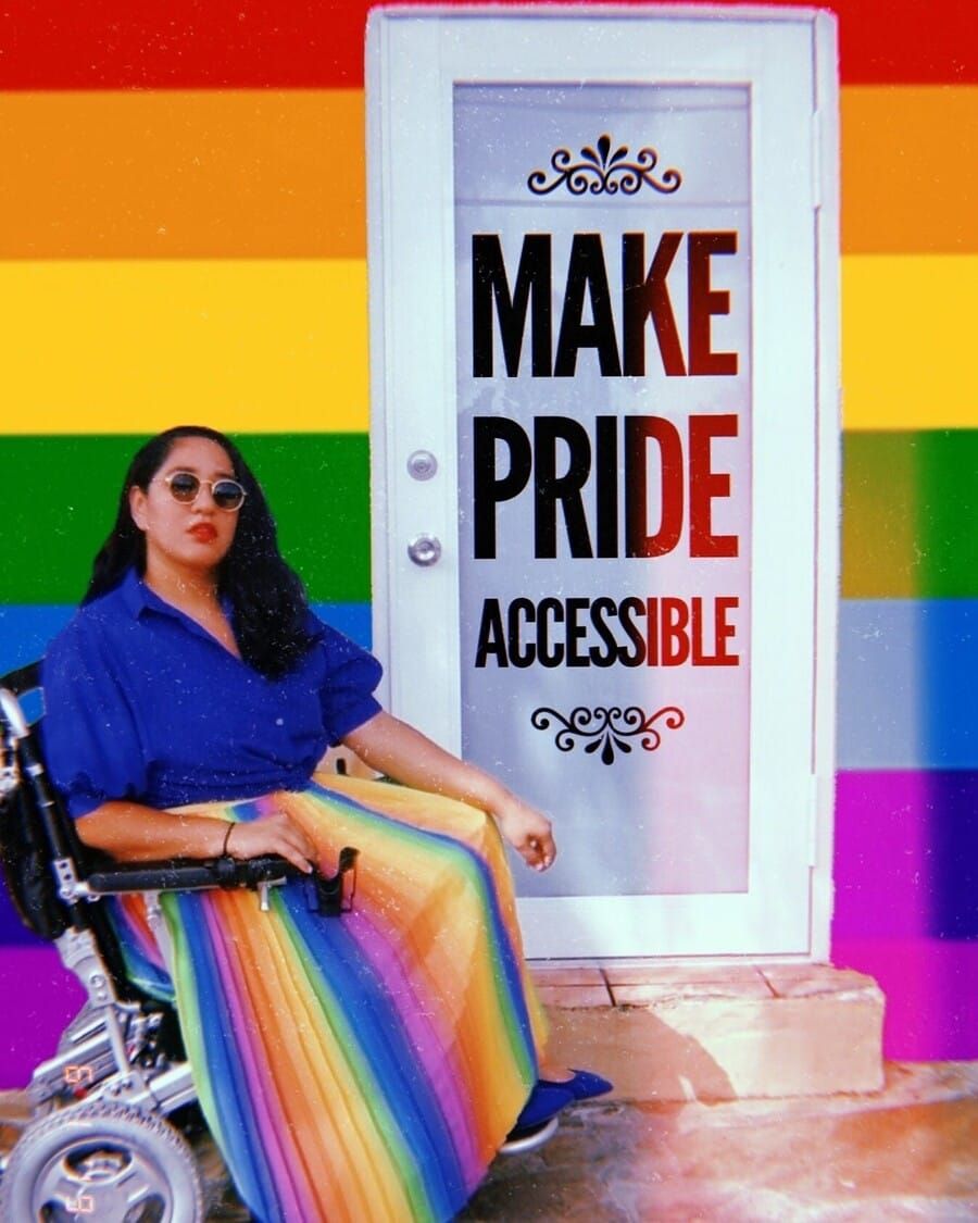 Annie Segarra, a Latinx American person, sits in a wheelchair wearing a rainbow skirt next to a doorstep and door that reads “Make Pride Accessible.” 