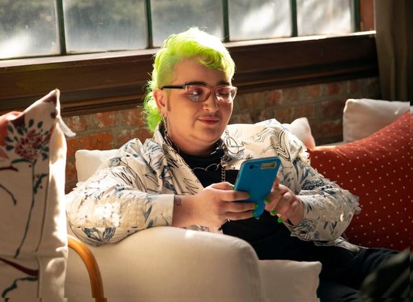A genderqueer person with neon hair sitting on a couch looking at their phone.
