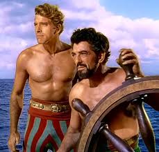 Burt Lancaster and Nick Cravat in a promotional still from The Crimson Pirate. They're both shirtless, and Cravat is gripping the wheel of a sailing ship.
