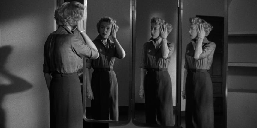 A still from the film Jennifer showing Ida Lupino, a slim woman with blond hair in a long pencil skirt and a blouse, looking at her three reflections in a three-fold mirror.
