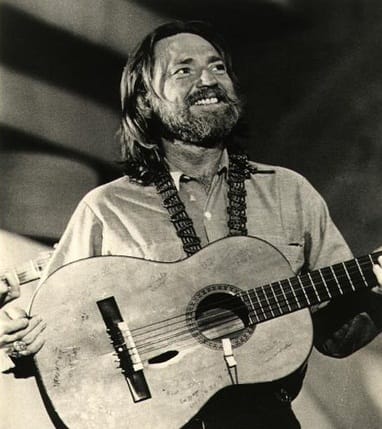 Willie Nelson at age 41 standing with his guitar on a strap around his neck. One hand is on the neck of the guitar, the other cradling its far end. He's smiling and looking away and up to his left. His hair is long and a bit unkempt, hanging to his shoulders, though his beard, while full, is nicely trimmed.