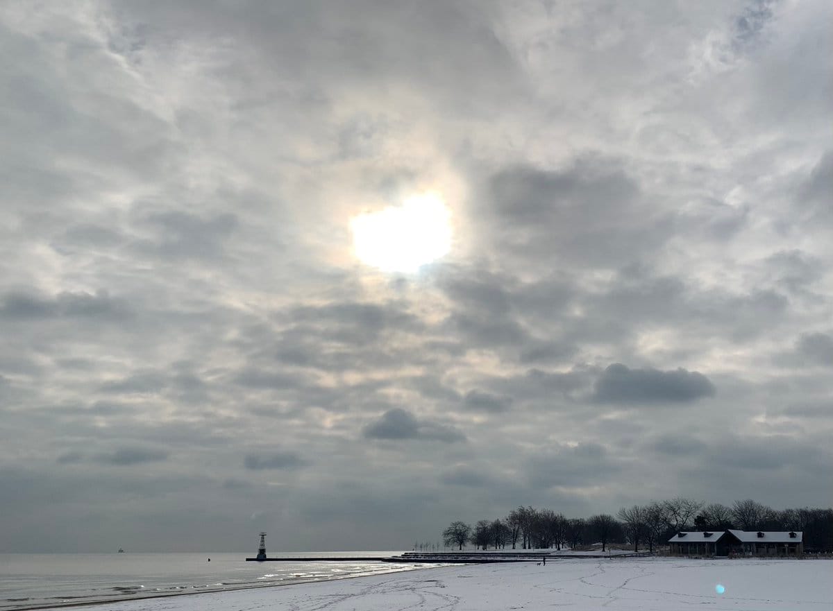 A photograph of Lake Michigan seen from Foster Beach, looking south. The beach has a modest covering of snow, and there is some ice along the water's edge. The sky above is full of clouds, with the sun shining through a hole in the center.