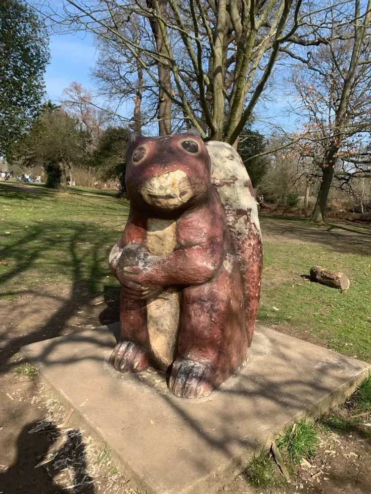 A photograph of a wooden carving of a squirrel, holding an acorn. It's about four feet tall and fairly crudely carved and painted. It's standing in a park.