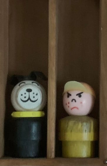 Two small wood-and-plastic action figures from the 1970s. One is of a dog with a black body and white head, the other is of a boy who is scowling and wears a yellow shirt and a ballcap with the brim tilted up.