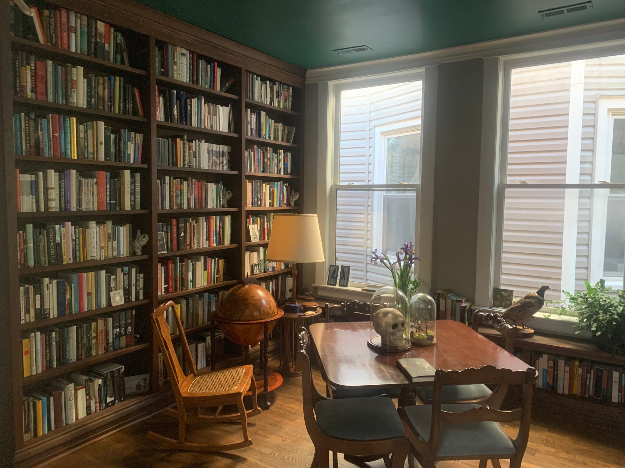 Our front library, with dark-stained built-in bookcases floor to ceiling on one wall and a low shelf below two large windows on another. A square table with chairs around it is in the center of the room on a hardwood floor, with a wooden rocking chair and a fake-antique globe behind them. A taxidermied pheasant stands on the windowsill.