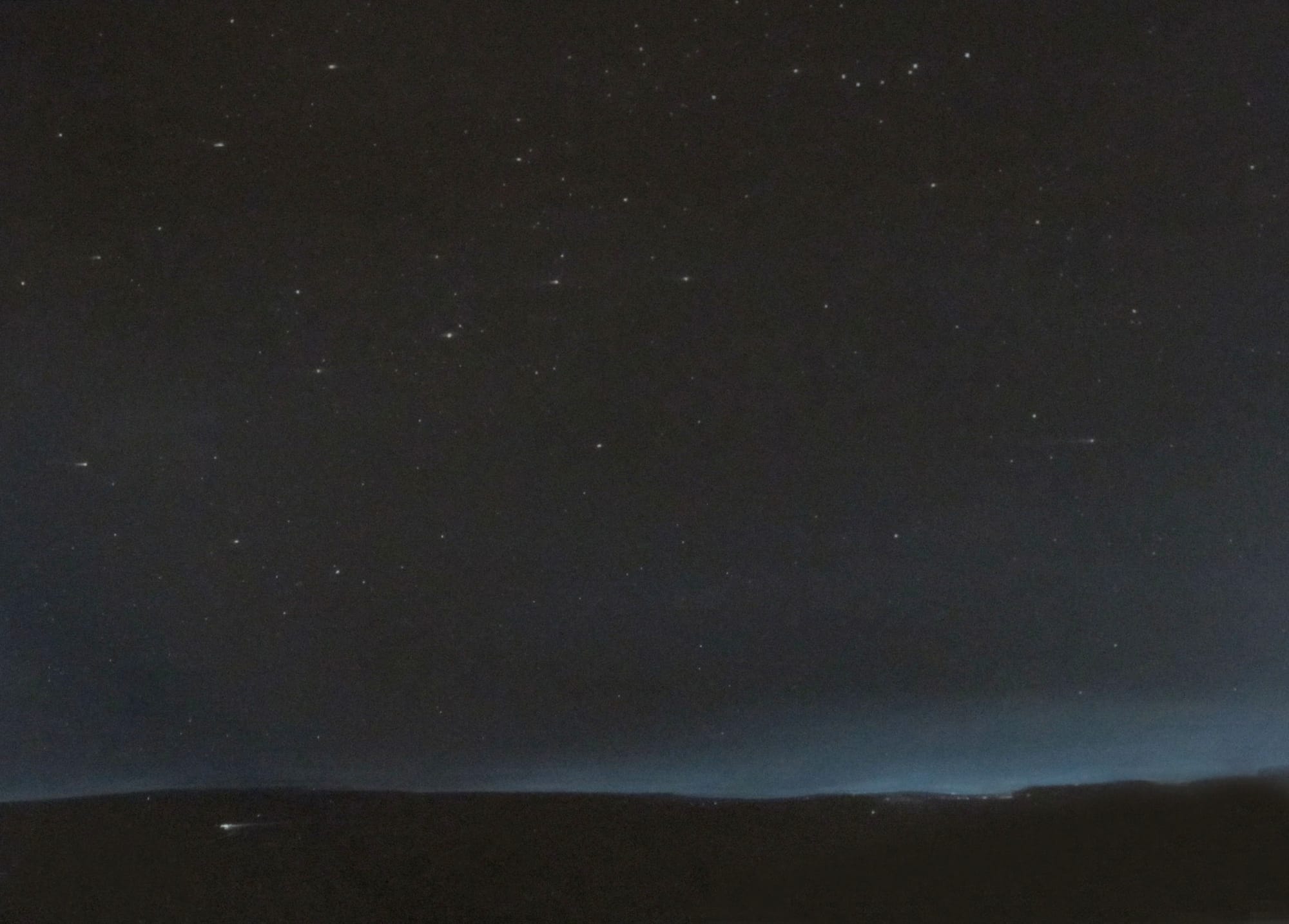 A canvas that is painted in shades of blue and black. The bottom is darkest, forming a low horizon, with above it a thin line of lighter blue-white, then above that an increasingly dark sky with, as you approach the top, some stars. Down in the landscape portion there are a couple of streaks of light, like headlights glimpsed form far off, and a few other fugitive light sources that are hard to make out.