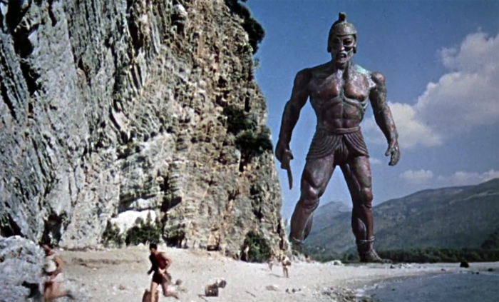 A still from Jason and the Argonauts showing some men running on a beach by a cliff, trying to get away from a giant, animated statue of a man with a sword.