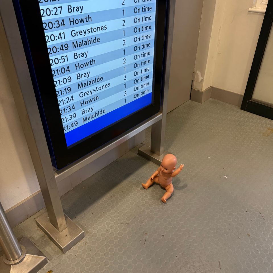 An abandoned toy doll (Naked), in front of a train departure screen. 