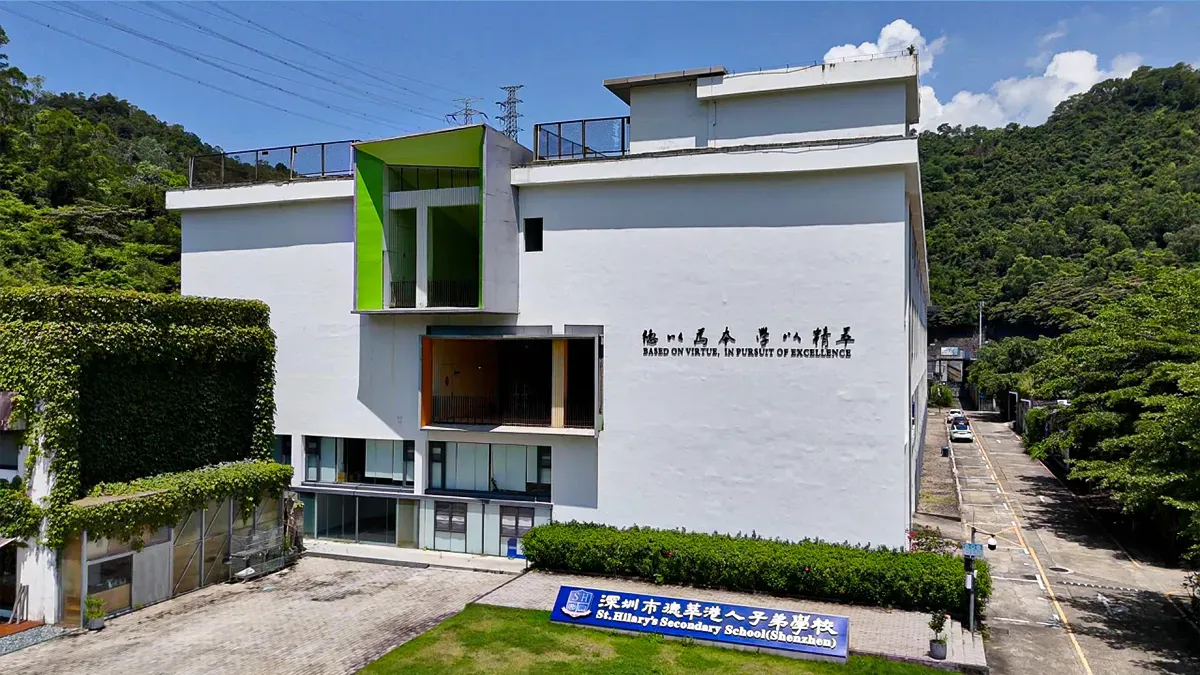 St. Hilary's Secondary School (Shenzhen) campus exterior — white building with green accent panels, school motto "Based on Virtue, In Pursuit of Excellence" on the facade, set against a wooded hillside
