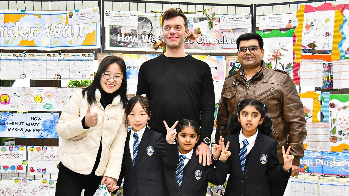International and Chinese families with students in Qingdao MINGDE School (QMDS) uniforms in front of an IB PYP inquiry display — bilingual school community, West Coast New Area, Qingdao