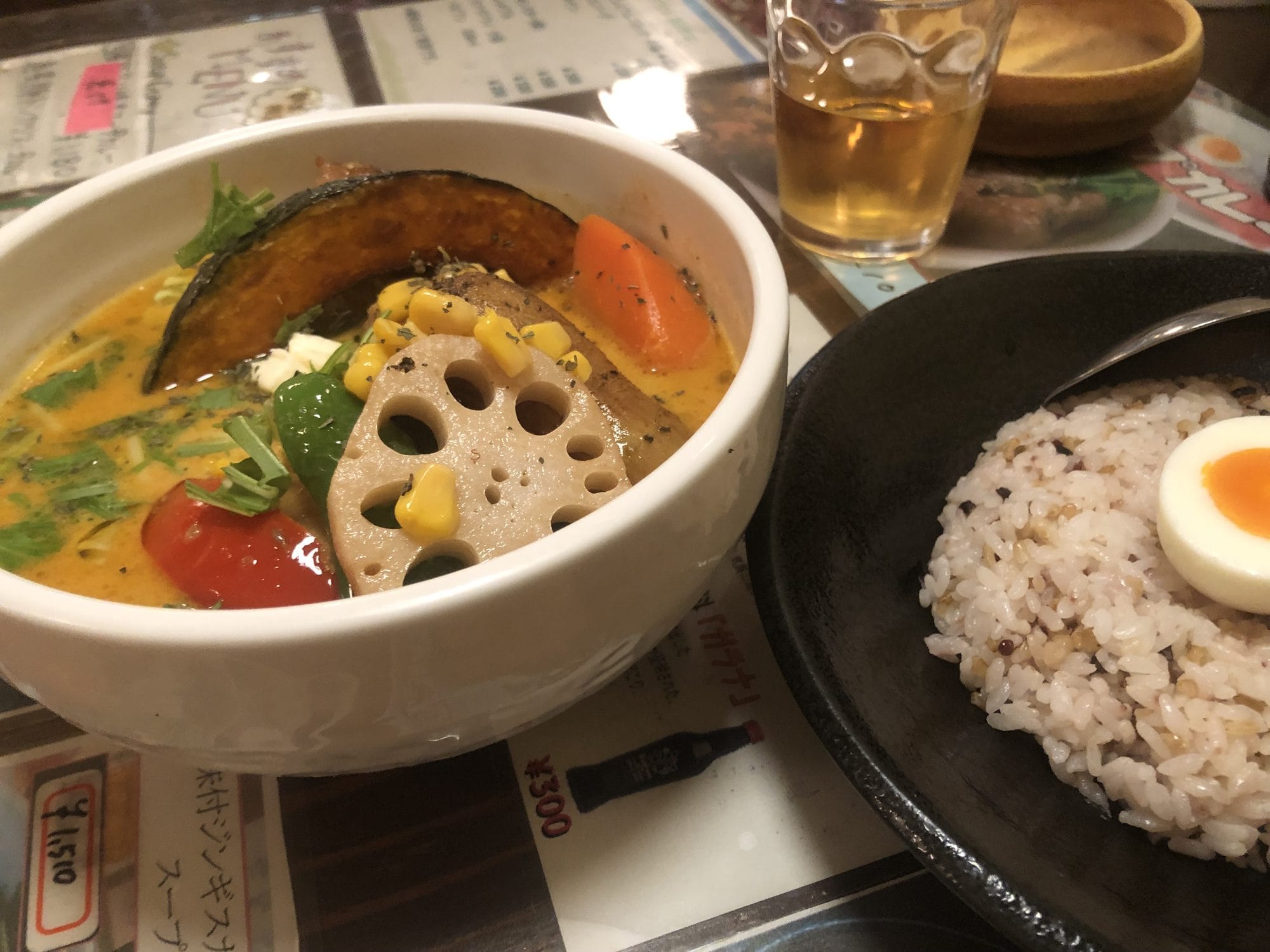 A bowl of Sapporo soup curry with pumpkin, lotus root, corn, and mixed vegetables, served with rice and a drink