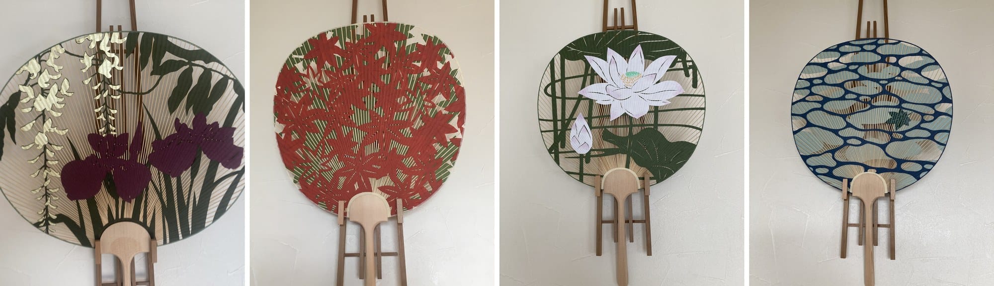 Four round Kyoto uchiwa by Aiba displayed on wooden stands, showing wisteria and iris, red maple leaves, a white lotus and a blue water pattern