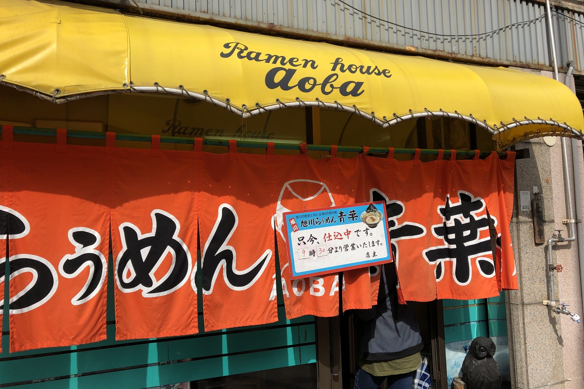 The shopfront of a long-established Asahikawa ramen shop, with a yellow awning reading Ramen house aoba and a red noren curtain