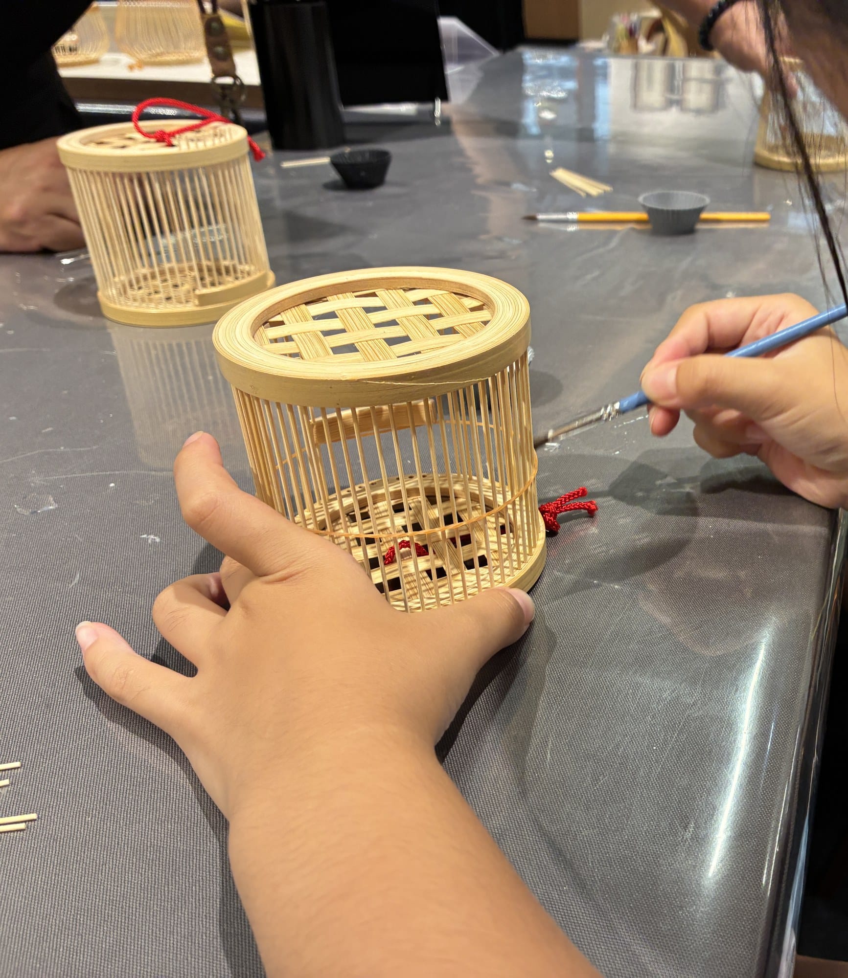 A child's hands gluing the frame of a bamboo insect cage at a workshop table at Japan Traditional Crafts Aoyama Square in Tokyo