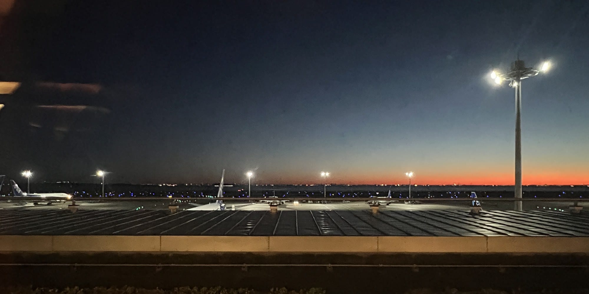 Haneda Airport before dawn, with aircraft parked on the apron under floodlights, blue taxiway lights and a thin orange band along the horizon