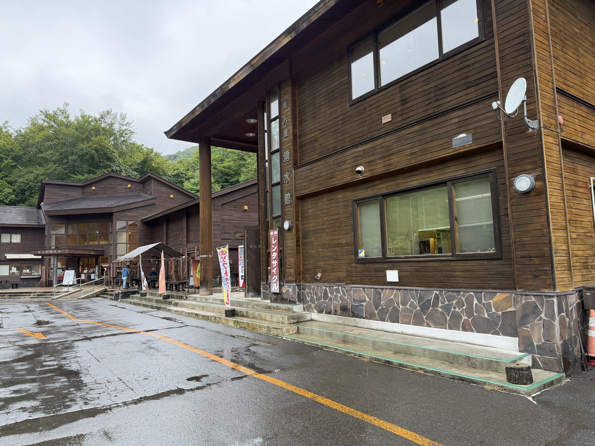 Exterior of the Oirase Keiryukan visitor building in Aomori on a rainy day, with a rental bicycle banner and car park