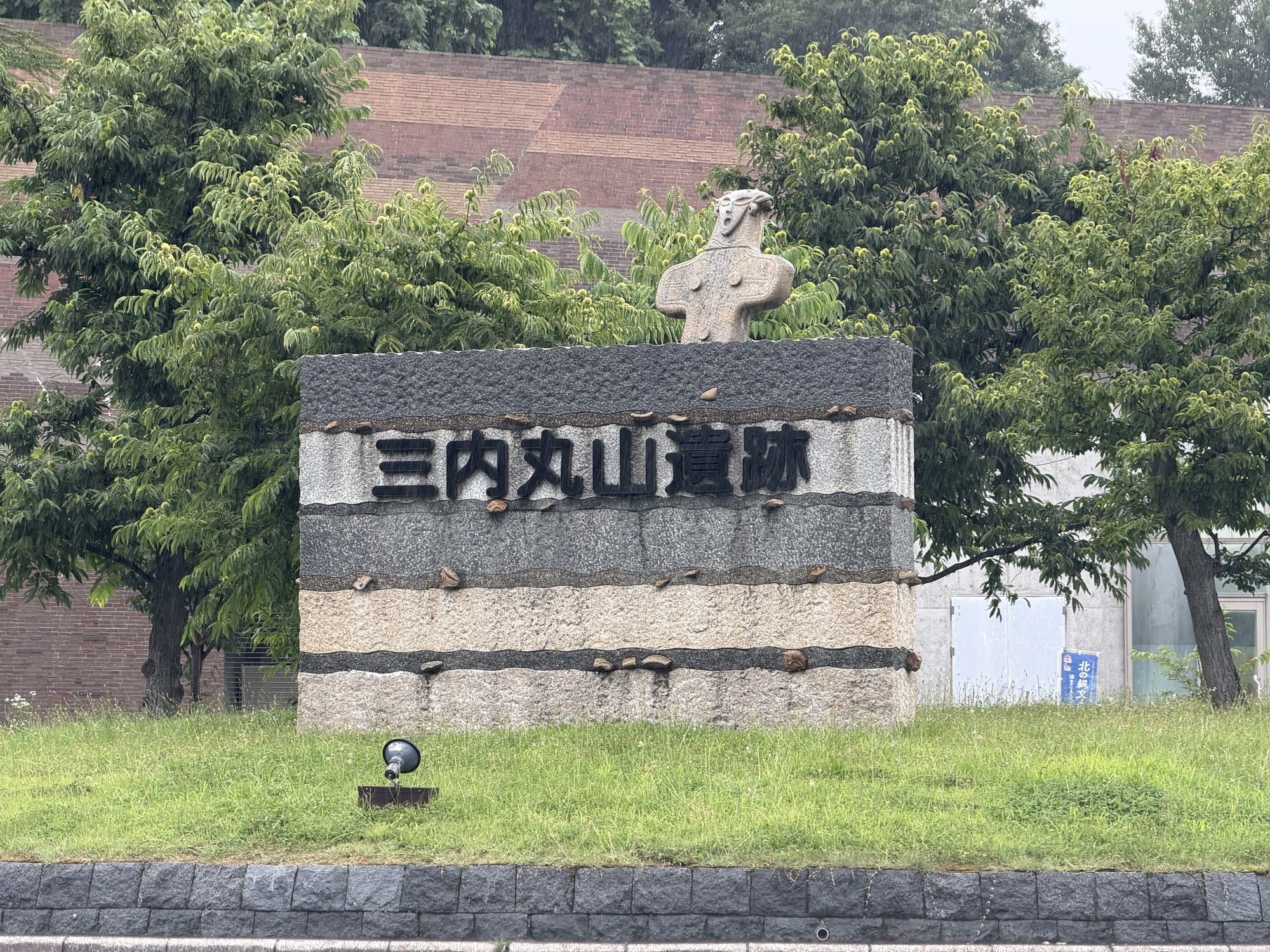 Stone marker reading Sannai-Maruyama Iseki at the site entrance in Aomori, topped by a carved figure shaped like a flat clay figurine, in the rain