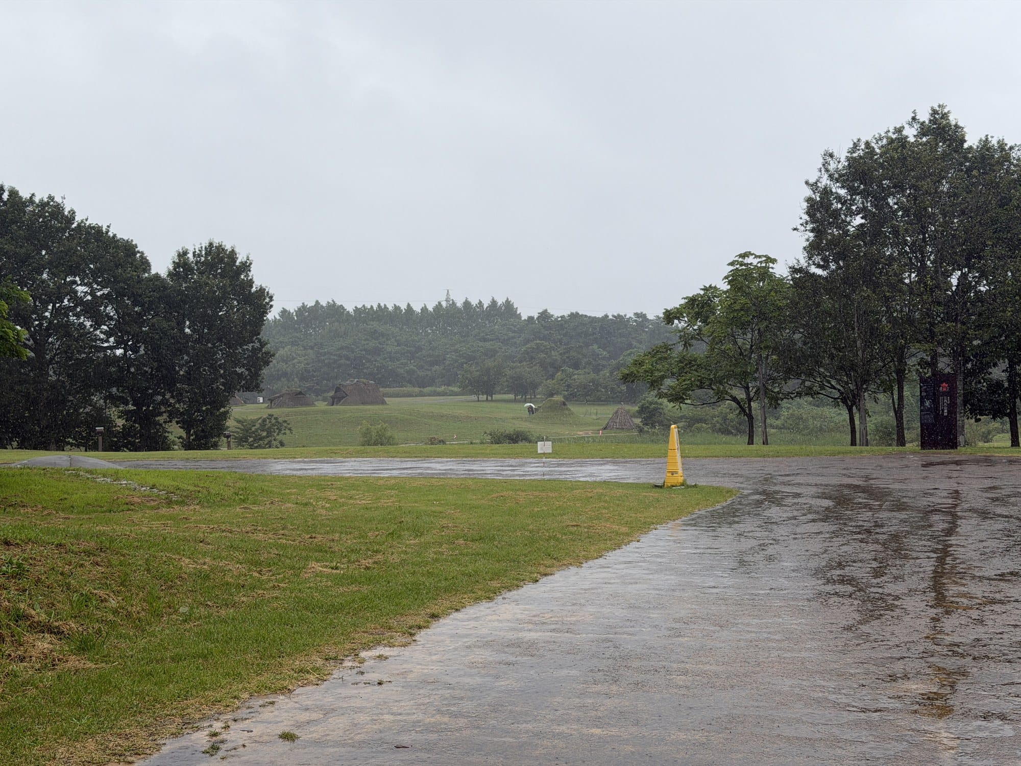Reconstructed thatched pit dwellings across open turf at the Sannai-Maruyama Jomon site in Aomori under heavy rain