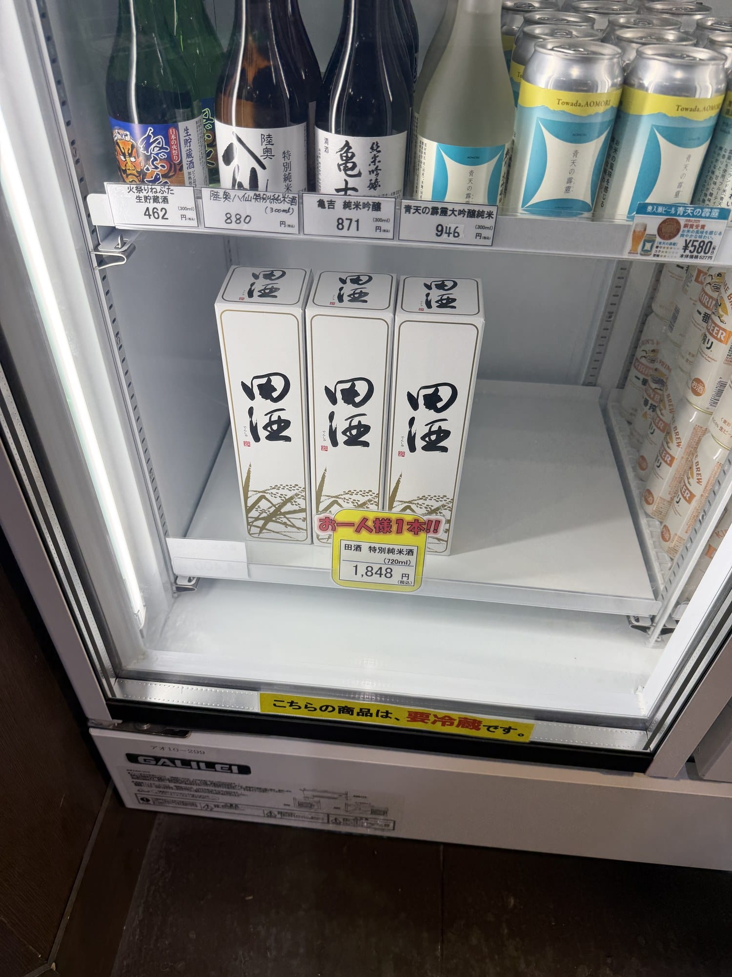 Refrigerated case at Shin-Aomori Station holding Denshu Tokubetsu Junmai with a one-bottle-per-customer sign, plus 300 ml Aomori sake and Oirase beer