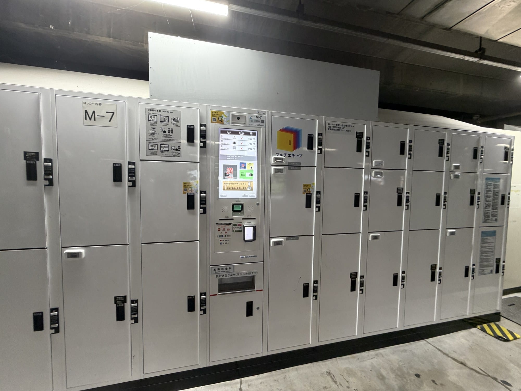 A long bank of grey coin lockers under a station deck, with a payment kiosk in the middle