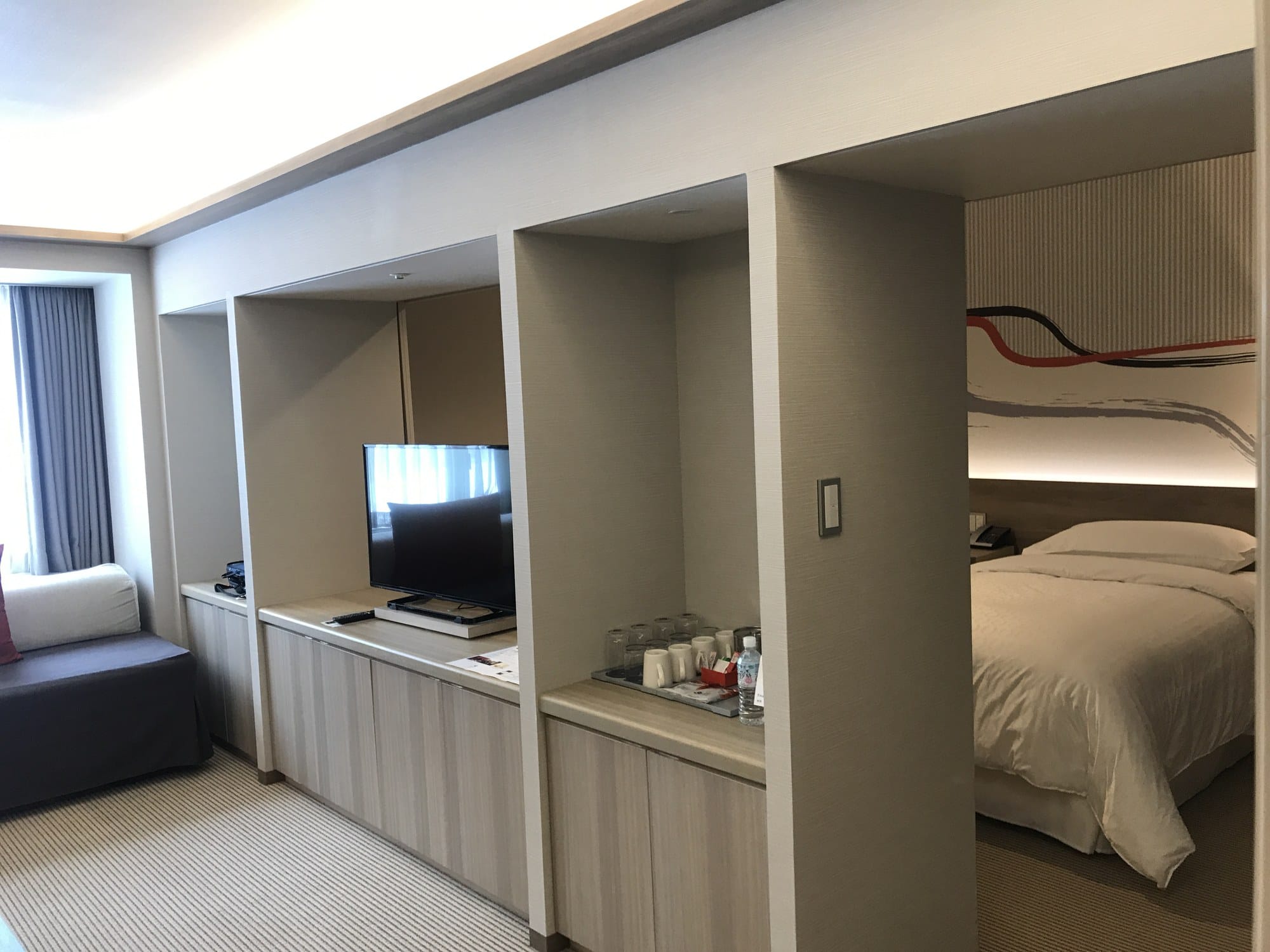 A hotel room divided by a partition wall, with a bed visible through the opening on the right and a sofa and television area in the foreground