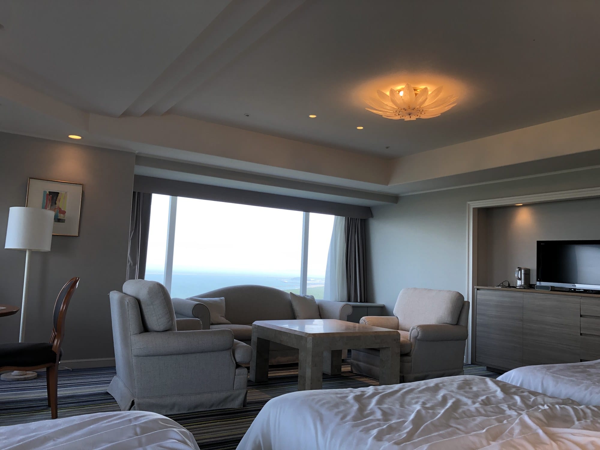 A large hotel room with two beds in the foreground, a separate seating area with armchairs and a sofa, and a wide window looking out to the sea