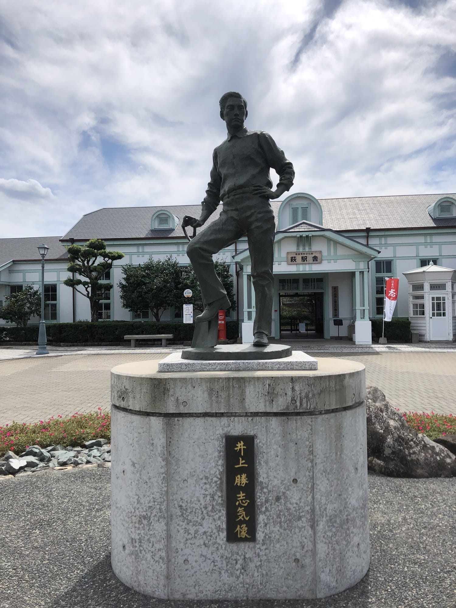 Bronze statue of a young Inoue Masaru with one foot on a shovel, in front of the white and pale-blue Western-style Hagi Station building in Yamaguchi Prefecture