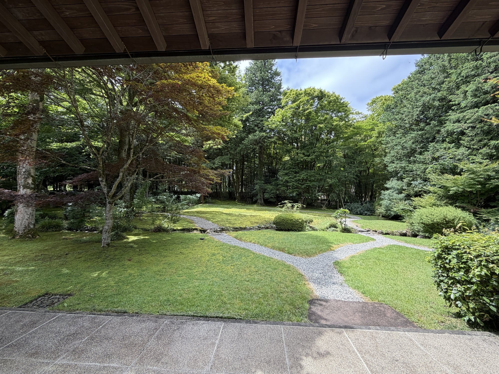 View from under the deep eaves of the Former Kishi Residence across a lawn where a gravel path forks in a Y shape, with maples and woodland behind
