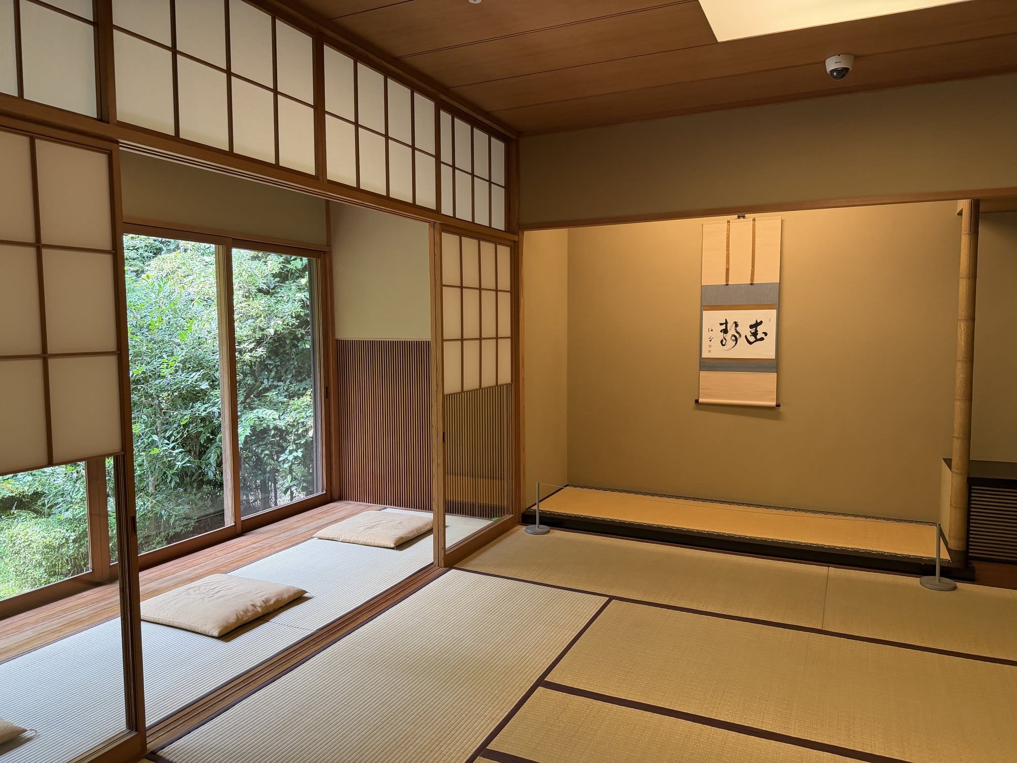 A tatami room in the Former Kishi Residence with a hanging scroll in the alcove, a bamboo post beside it, shoji screens and two floor cushions on the veranda