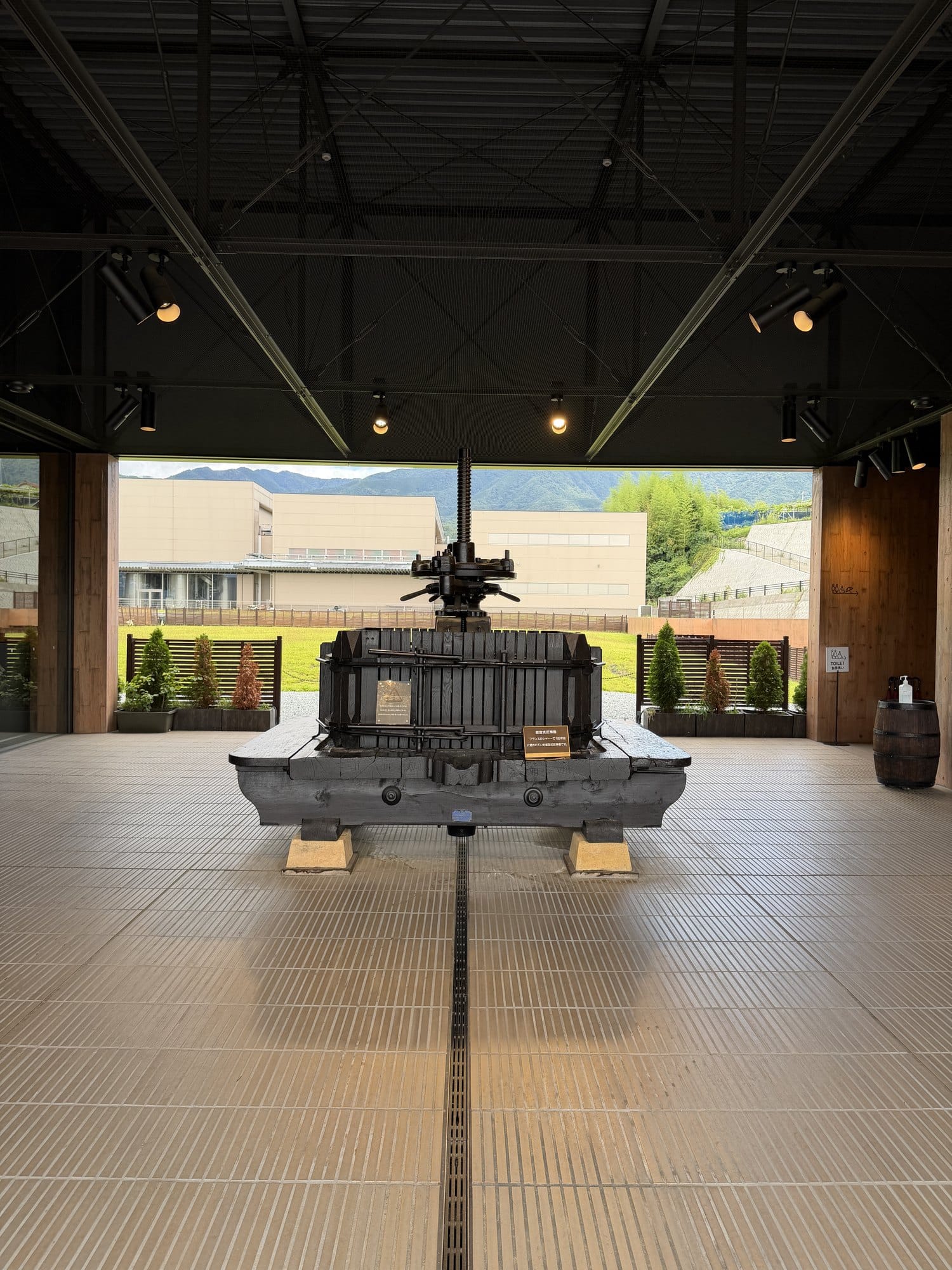 An old wooden and iron grape press displayed in a hall at Mannns Wines Katsunuma, with the hills visible through the open end
