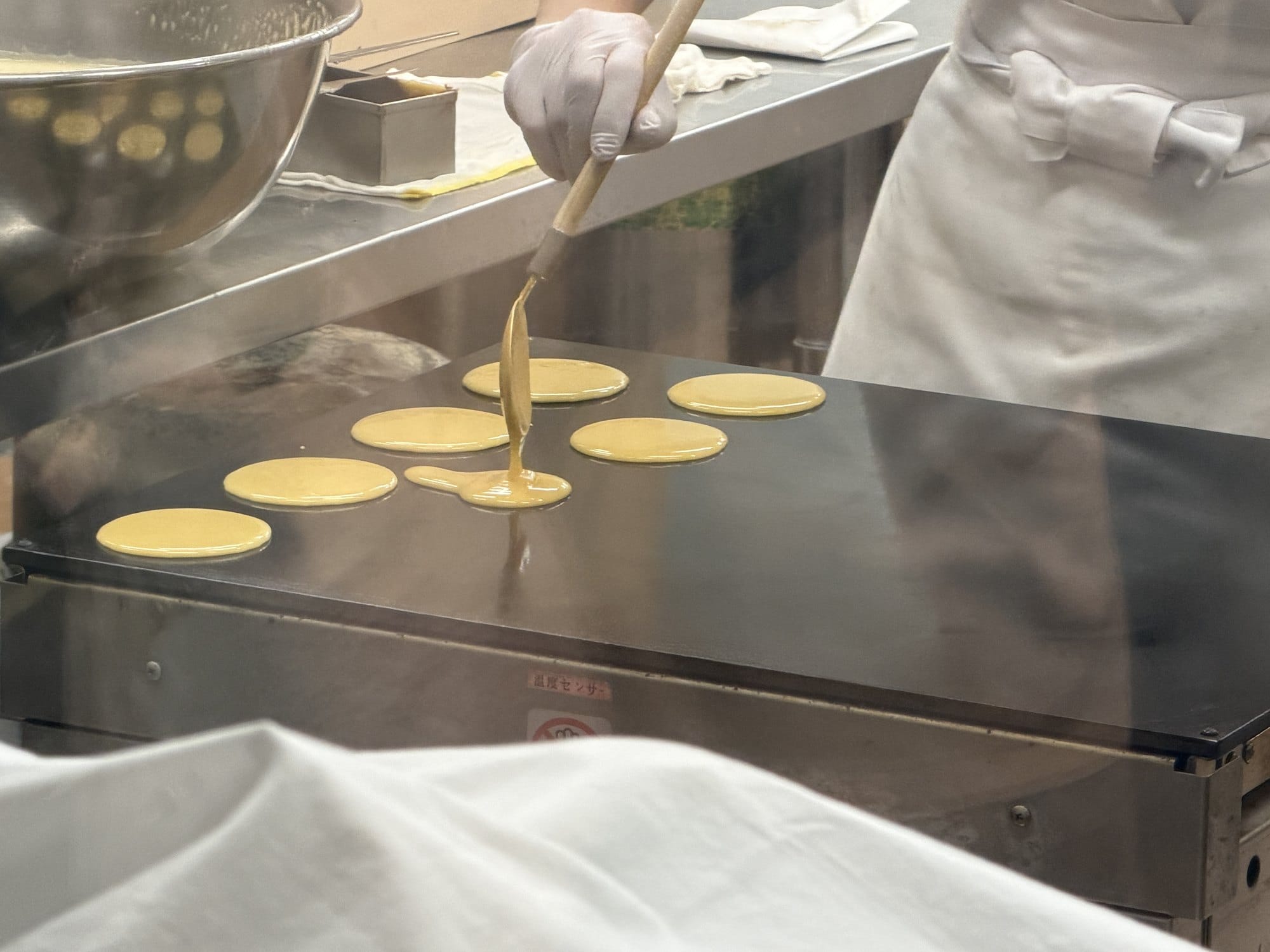 Batter poured from a wooden ladle onto a wide hot plate at Toraya Kobo, six rounds of dorayaki cooking at once