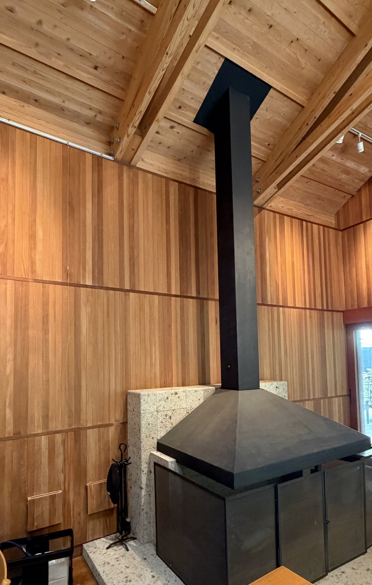 Inside Toraya Kobo: cedar-board walls, exposed rafters and a black steel stove with a square flue rising to the roof, on a pale stone hearth